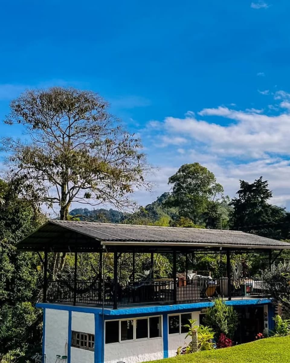 Edificio blanco y azul de dos niveles con terraza cubierta en el piso de arriba, prado verde, árboles altos y una colina al fondo bajo un cielo azul con nubes.