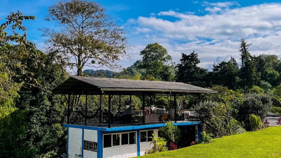 Edificio blanco y azul de dos niveles con terraza cubierta en el piso de arriba, prado verde, árboles altos y una colina al fondo bajo un cielo azul con nubes.