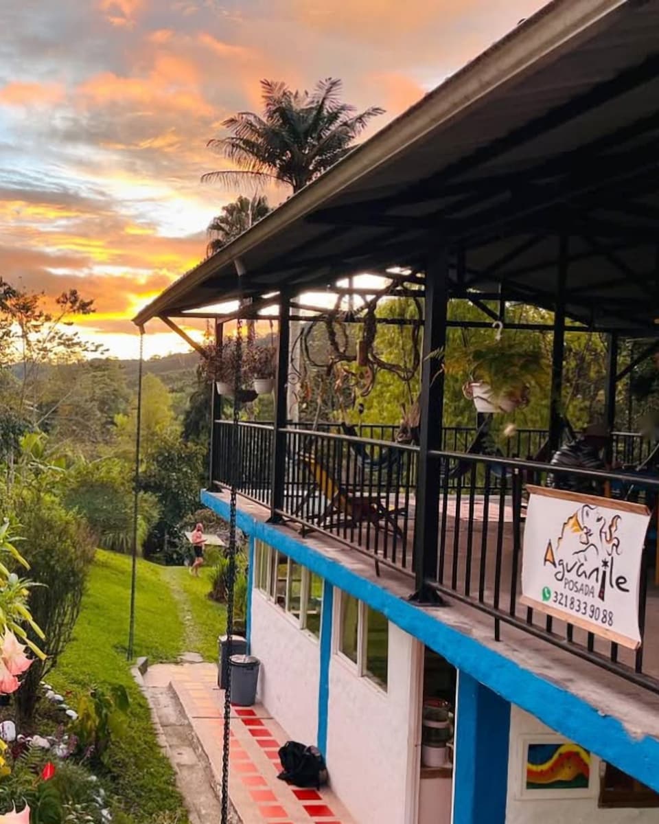La casa principal al atardecer: balcón con baranda negra, borde azul y el letrero pintado a mano de Aguanile, con flores de borrachero color durazno en primer plano y cielo naranja.