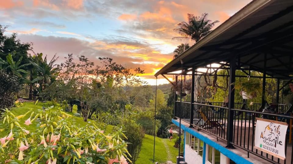 La casa principal al atardecer: balcón con baranda negra, borde azul y el letrero pintado a mano de Aguanile, con flores de borrachero color durazno en primer plano y cielo naranja.