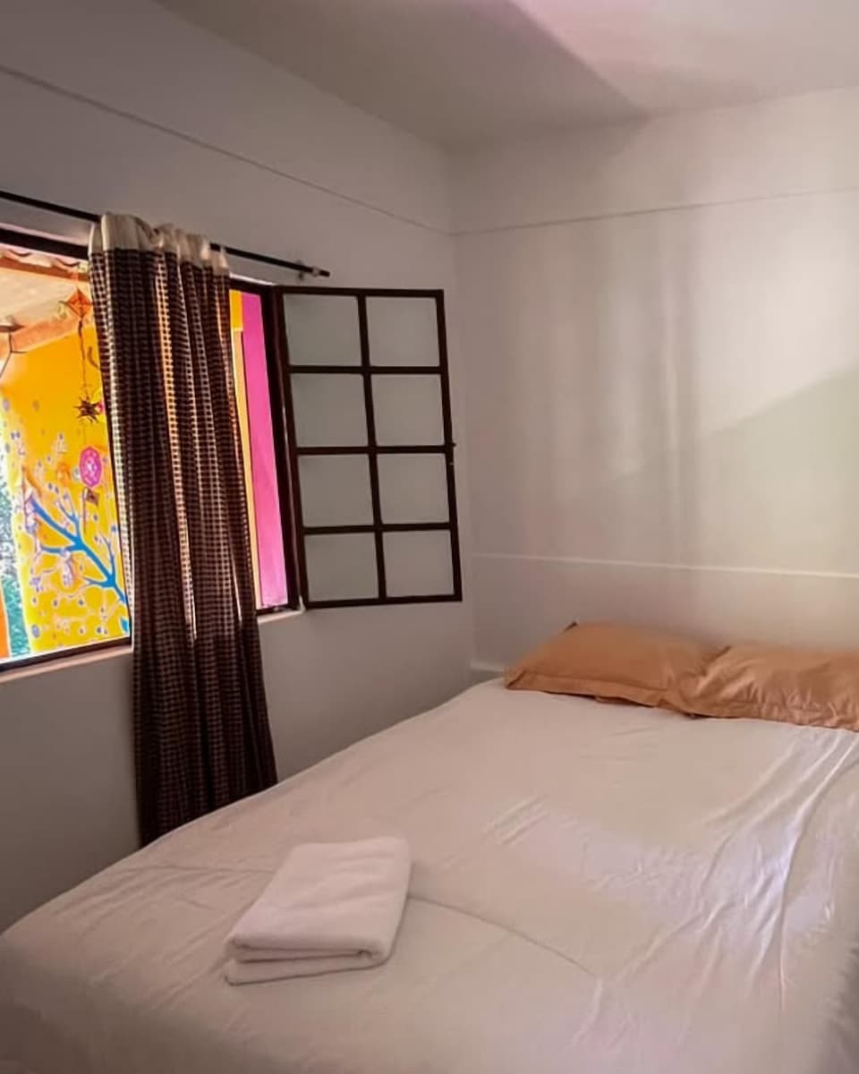Habitación privada con cama doble de sábanas blancas, almohadas color durazno, una toalla doblada y una ventana de madera que deja ver una pared pintada de amarillo.