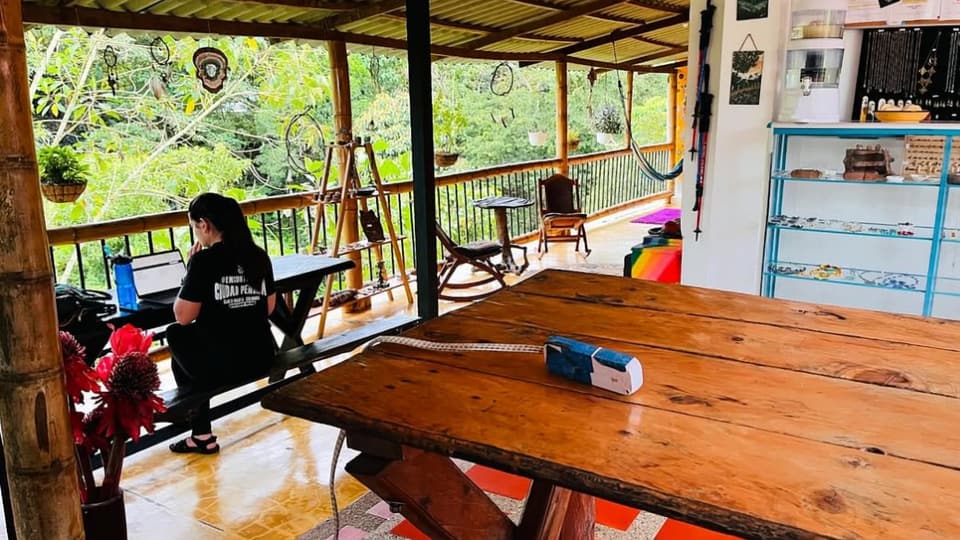 Mesa larga de madera rústica en la terraza cubierta, piso de baldosas rojas, una persona vista de lado usando un portátil junto a la baranda y, al fondo, una hamaca y un estante con artesanías.